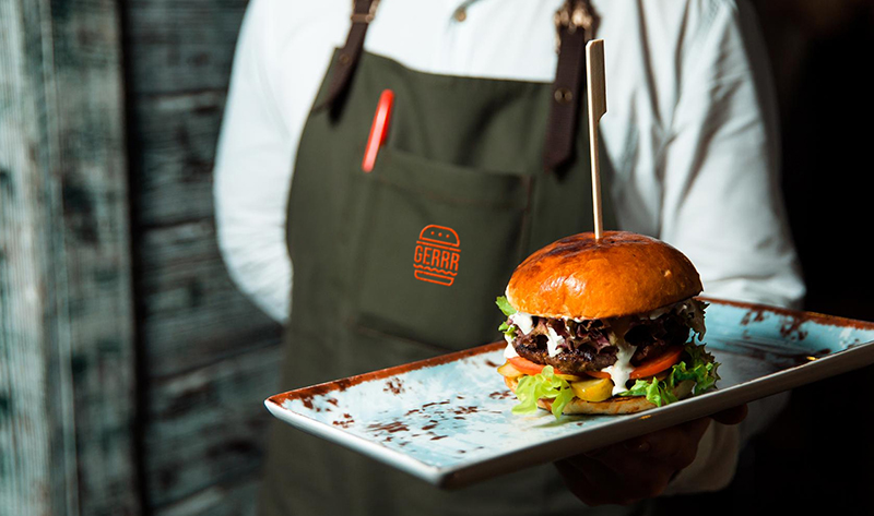 A server in a GERRR Burger apron holding a plate with a gourmet burger garnished with lettuce, tomato, and a skewer through the bun.